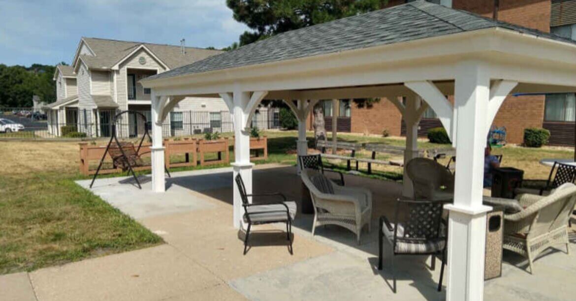 A white outdoor pavilion with chairs and a swing beside grass and apartment buildings under a blue sky.