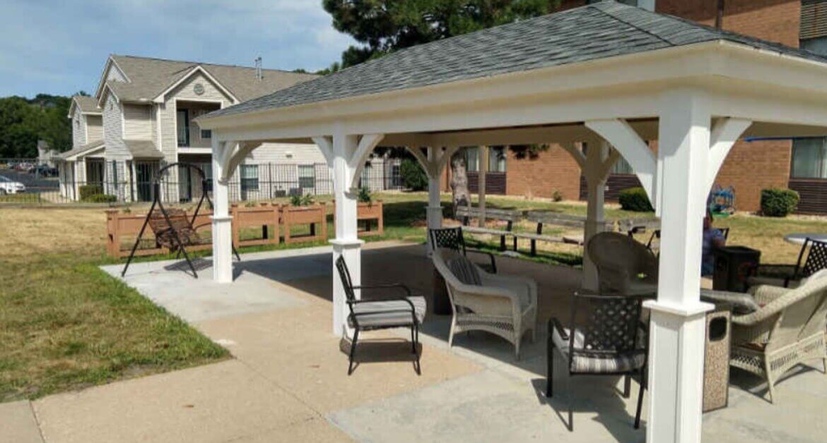 A white outdoor pavilion with chairs and a swing beside grass and apartment buildings under a blue sky.