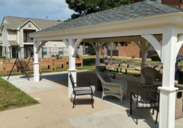 A white outdoor pavilion with chairs and a swing beside grass and apartment buildings under a blue sky.