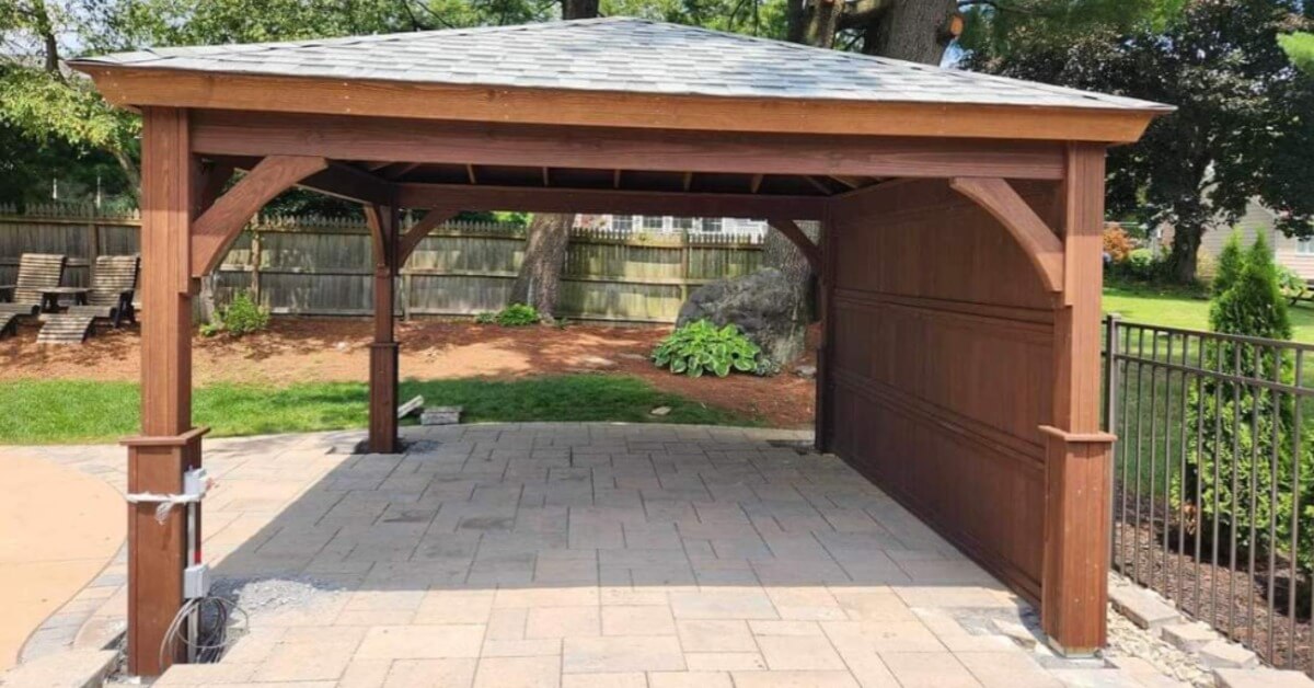 A wooden outdoor pavilion with a shingled roof standing over a paved patio near fencing and green trees.