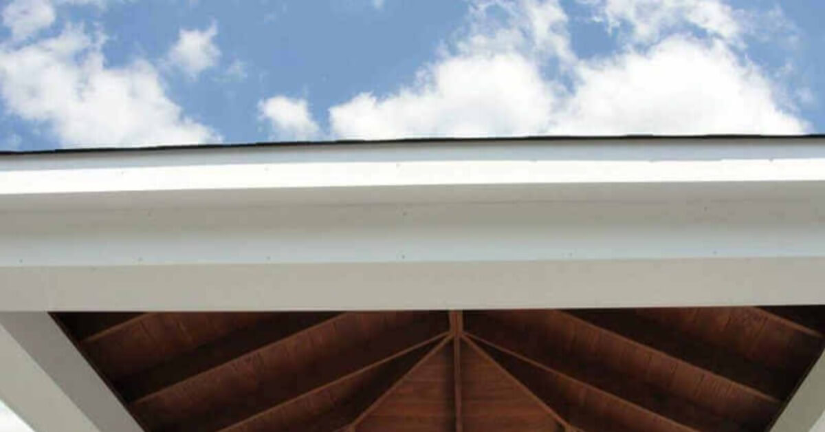 A close view of a white pavilion roof edge and dark wooden rafters beneath a blue sky with scattered clouds.