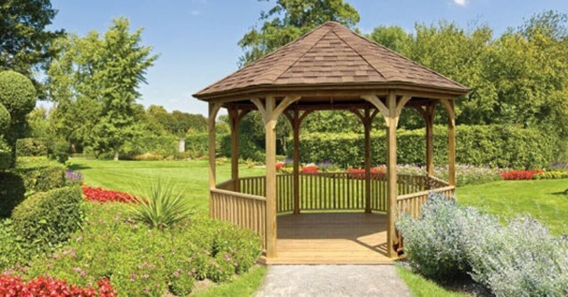 A wooden gazebo in a landscaped garden with trimmed hedges, flower beds, green lawn, and trees behind it.