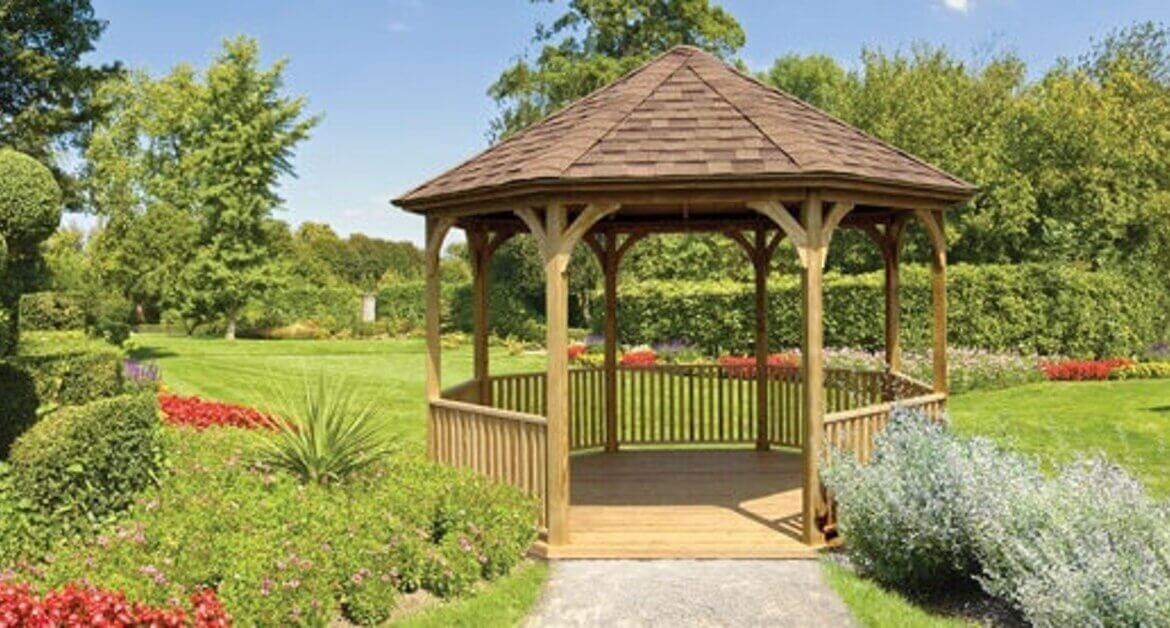 A wooden gazebo in a landscaped garden with trimmed hedges, flower beds, green lawn, and trees behind it.