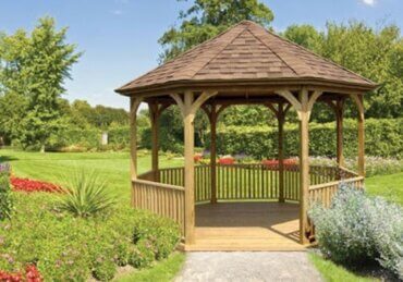 A wooden gazebo in a landscaped garden with trimmed hedges, flower beds, green lawn, and trees behind it.