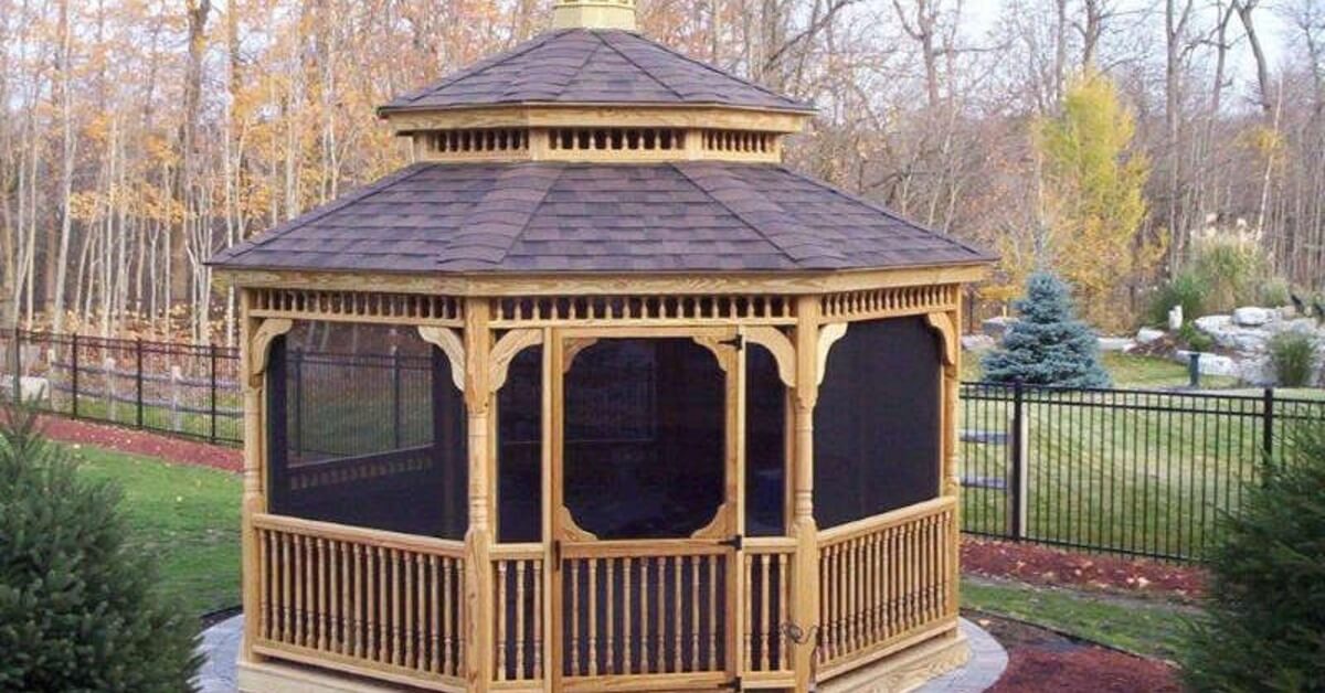 A screened wooden gazebo with a raised roof in a yard bordered by trees, fencing, shrubs, rocks, and mulch.