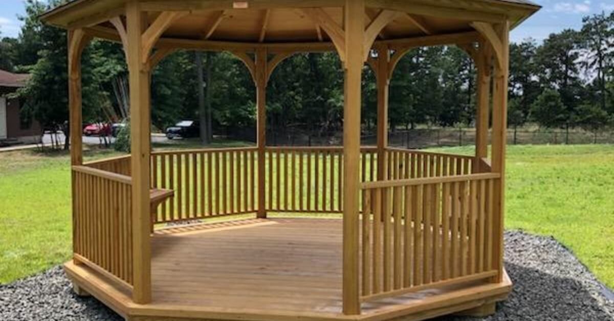 A wooden gazebo standing on a gravel base in a grassy yard with trees, vehicles, and a partly cloudy sky.