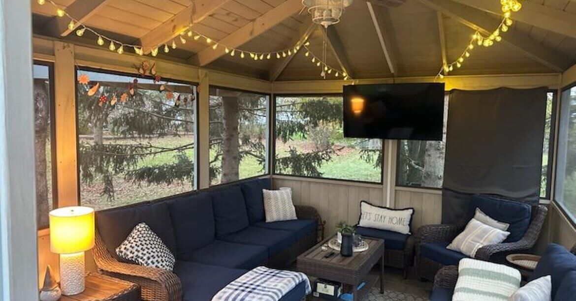 a screened gazebo with wicker seating, string lights, a ceiling fan, wall-mounted television, and trees outside.