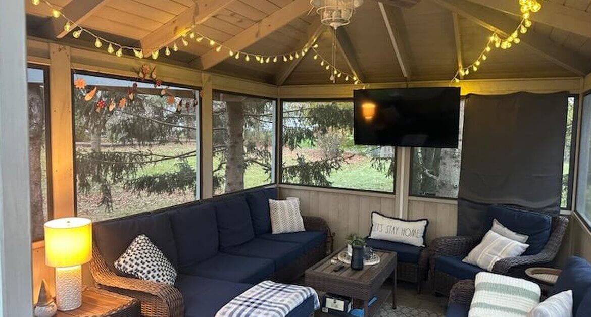 a screened gazebo with wicker seating, string lights, a ceiling fan, wall-mounted television, and trees outside.