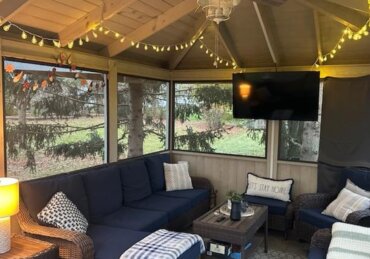 a screened gazebo with wicker seating, string lights, a ceiling fan, wall-mounted television, and trees outside.