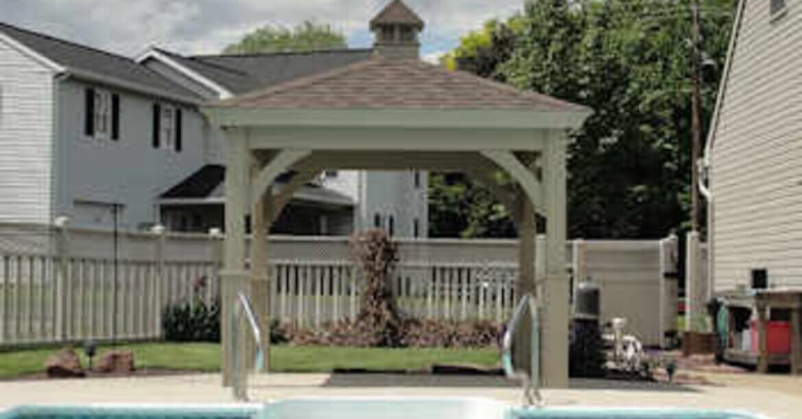 A white gazebo beside a backyard pool with a shingled roof, cupola, surrounding fence, homes, and trees.