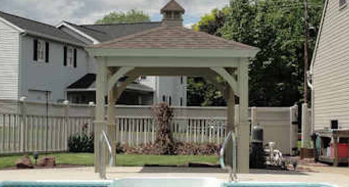 A white gazebo beside a backyard pool with a shingled roof, cupola, surrounding fence, homes, and trees.