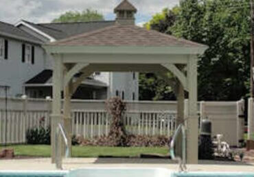 A white gazebo beside a backyard pool with a shingled roof, cupola, surrounding fence, homes, and trees.