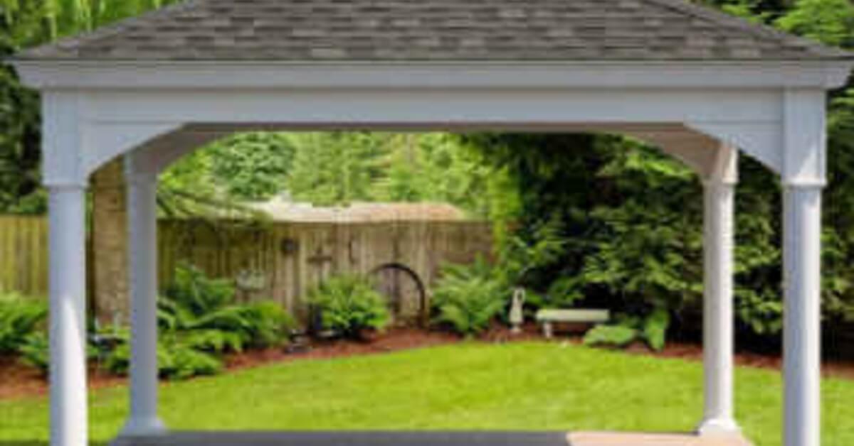 A white pavilion with a gray shingled roof standing on a backyard lawn near shrubs, trees, and a paved pad.