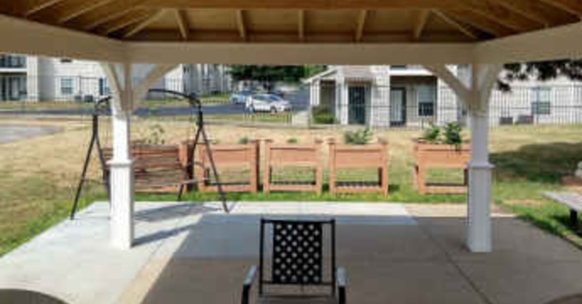 A covered pavilion with exposed wood beams, white posts, patio chairs, a trash can, and a yard beyond.