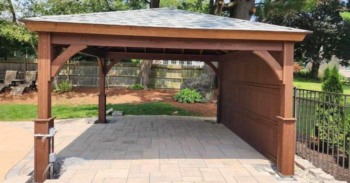 A wooden backyard pavilion with a shingled roof sits on a paved patio, surrounded by fencing and trees.