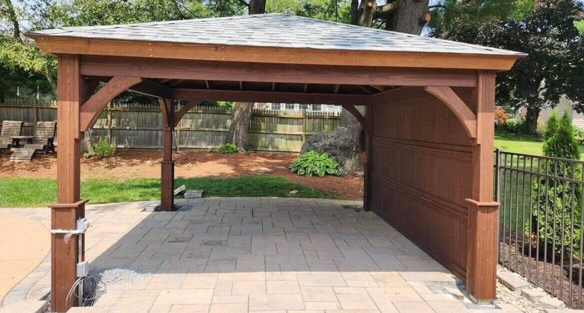 A wooden backyard pavilion with a shingled roof sits on a paved patio, surrounded by fencing and trees.