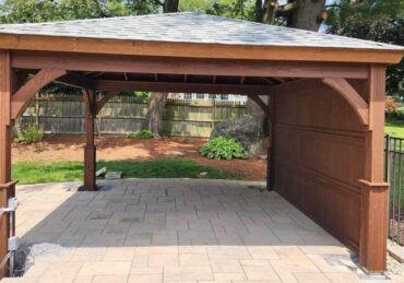 A wooden backyard pavilion with a shingled roof sits on a paved patio, surrounded by fencing and trees.