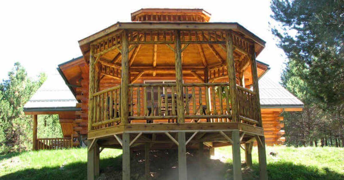 A raised natural wooden gazebo with railings sits on a grassy slope near a cabin, surrounded by tall trees.