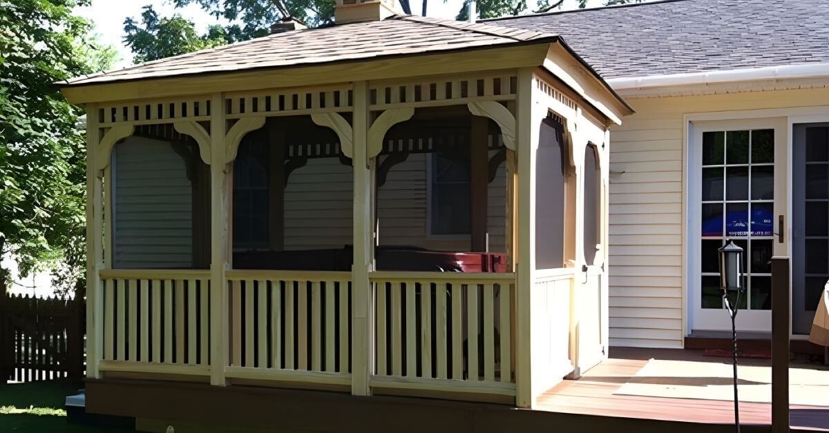 A screened wooden gazebo sits on a backyard deck beside a house, featuring railings, trim details, and a shingled roof.