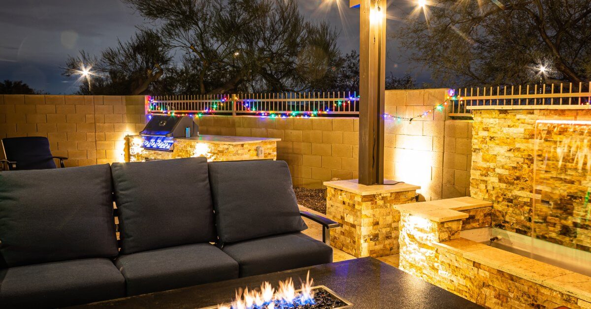 A wooden pavilion with a gas fire pit and cushioned seating stands beside a swimming pool in a landscaped backyard.