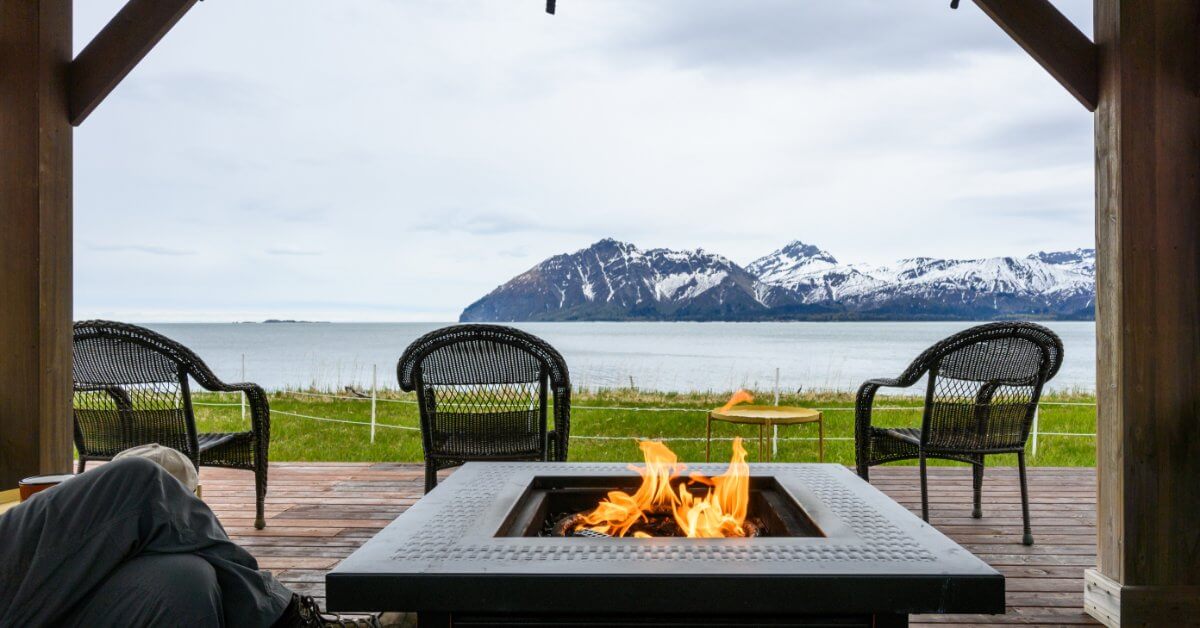 A lit fire pit with surrounding chairs sits beneath a covered wooden structure overlooking a lake and snowy mountains.