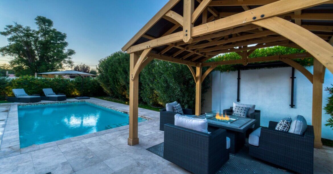 A lit fire pit with surrounding chairs sits beneath a covered wooden structure overlooking a lake and snowy mountains.