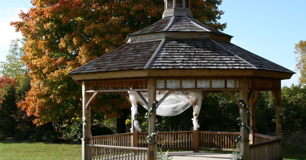 A wooden gazebo with flowing white curtains stands against colorful autumn trees in a grassy outdoor setting.