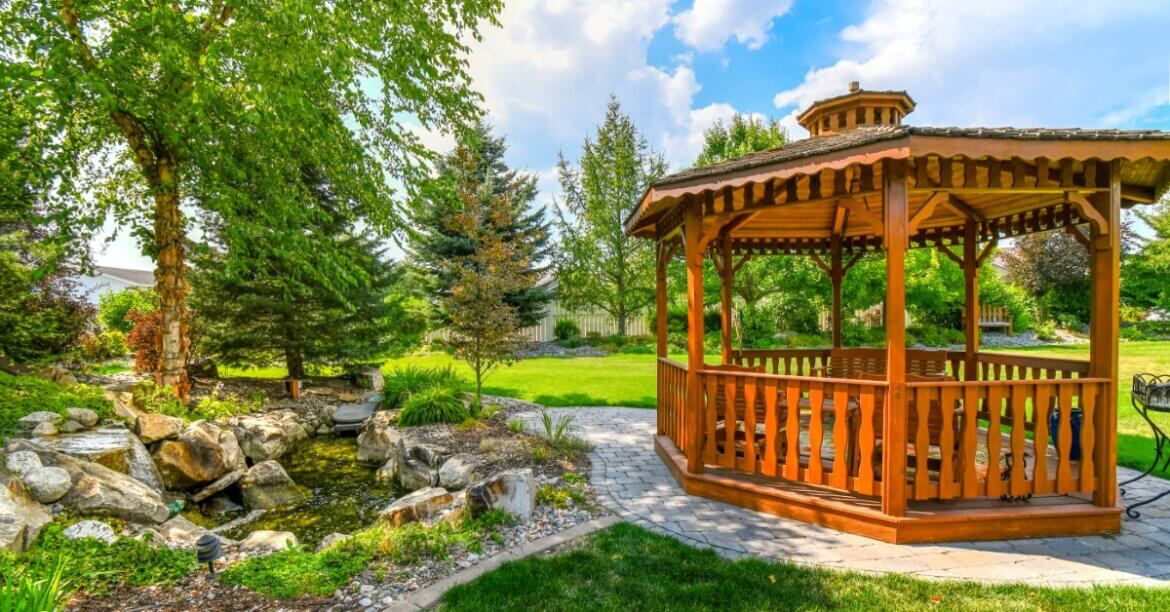 A wooden gazebo stands beside a landscaped backyard pond surrounded by mature trees, shrubs, and green lawn.