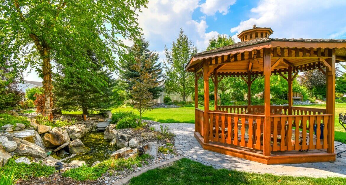 A wooden gazebo stands beside a landscaped backyard pond surrounded by mature trees, shrubs, and green lawn.