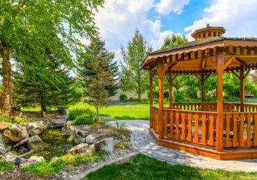 A wooden gazebo stands beside a landscaped backyard pond surrounded by mature trees, shrubs, and green lawn.