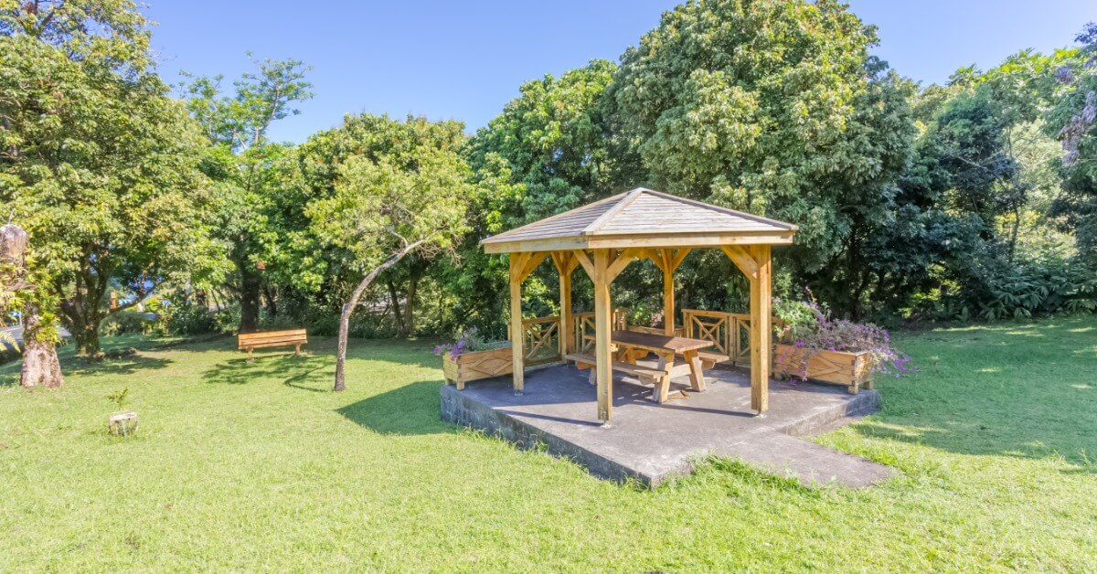 A wooden gazebo with open posts and picnic seating stands on a raised concrete foundation in a spacious green park.