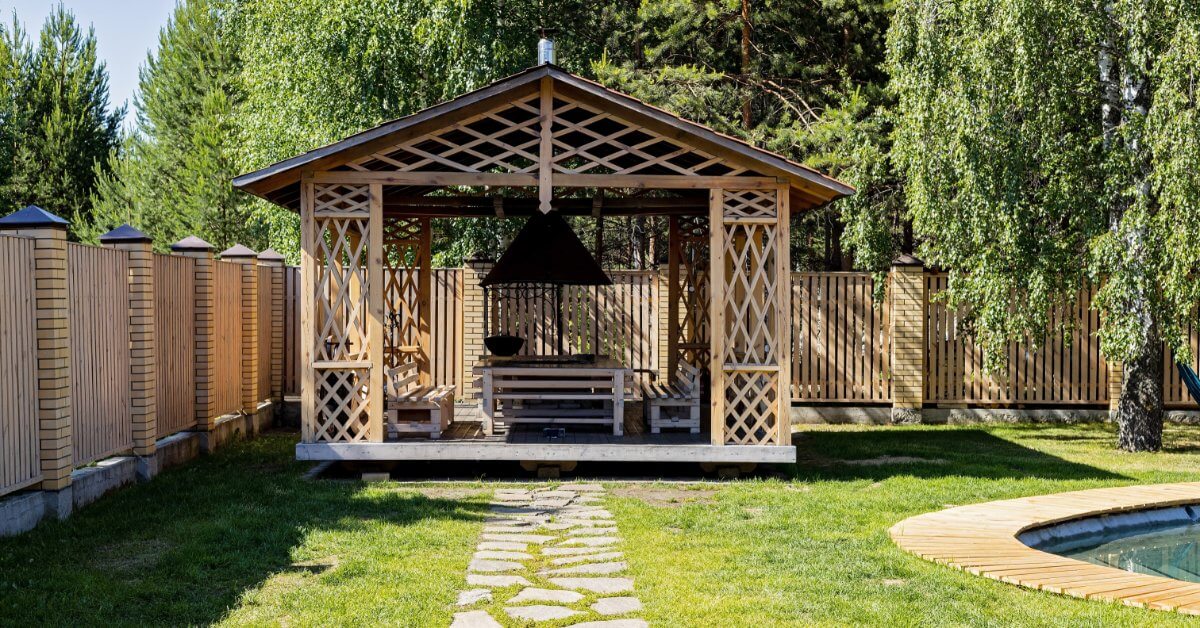A wooden gazebo with built-in seating stands on a lawn beside a swimming pool and stone garden path.