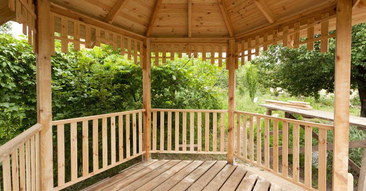 The interior of a wooden gazebo features exposed roof beams, railings, and natural wood flooring surrounded by greenery.