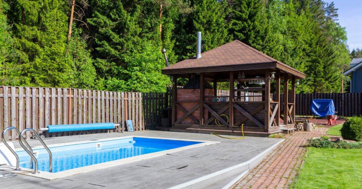 A wooden gazebo stands beside a backyard swimming pool with a paved deck, green lawn, and tall trees.