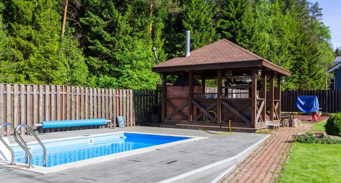 A wooden gazebo stands beside a backyard swimming pool with a paved deck, green lawn, and tall trees.
