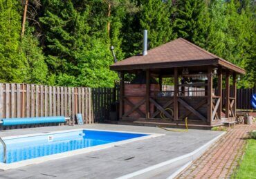 A wooden gazebo stands beside a backyard swimming pool with a paved deck, green lawn, and tall trees.