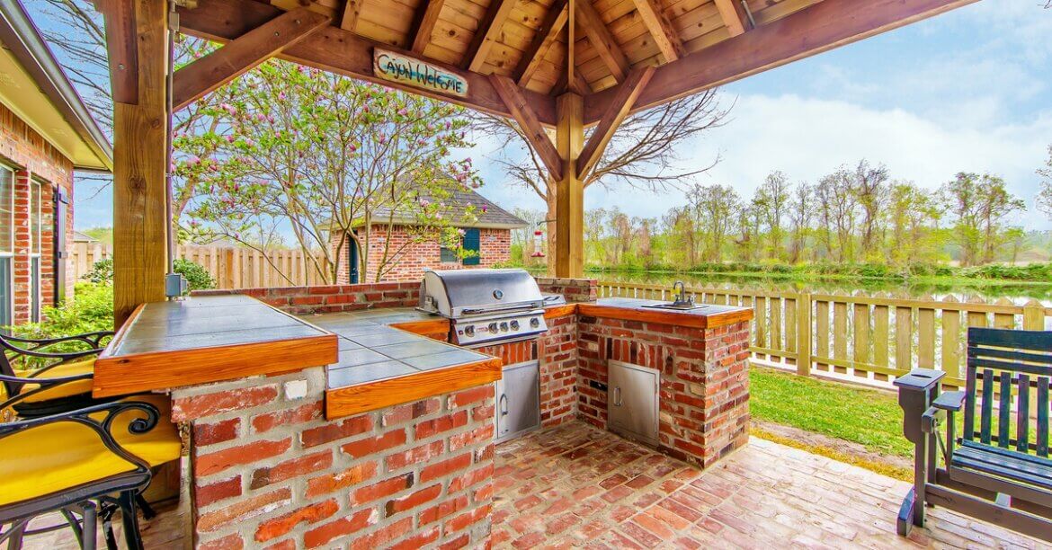 A wooden gazebo covers an outdoor kitchen with brick counters, a built-in grill, sink, and nearby seating.