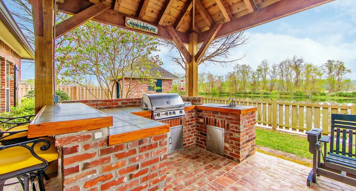 A wooden gazebo covers an outdoor kitchen with brick counters, a built-in grill, sink, and nearby seating.