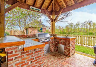 A wooden gazebo covers an outdoor kitchen with brick counters, a built-in grill, sink, and nearby seating.