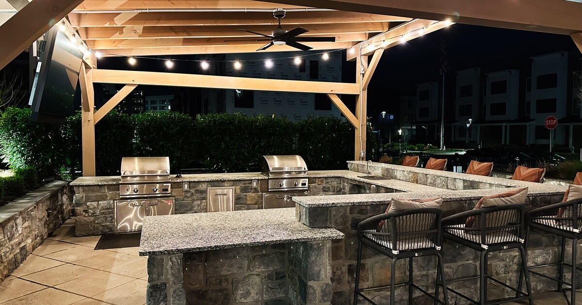 A covered outdoor kitchen features stone counters, built-in grills, bar seating, and overhead string lights.