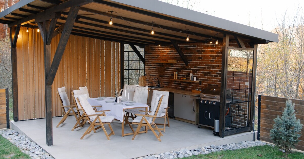 A covered outdoor kitchen combines a dining table and chairs with a grill, counter, sink, and brick wall.