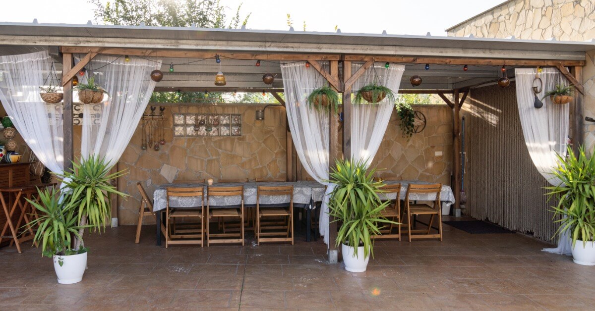 A covered outdoor dining area features hanging lights, long dining tables, wooden chairs, and potted plants.