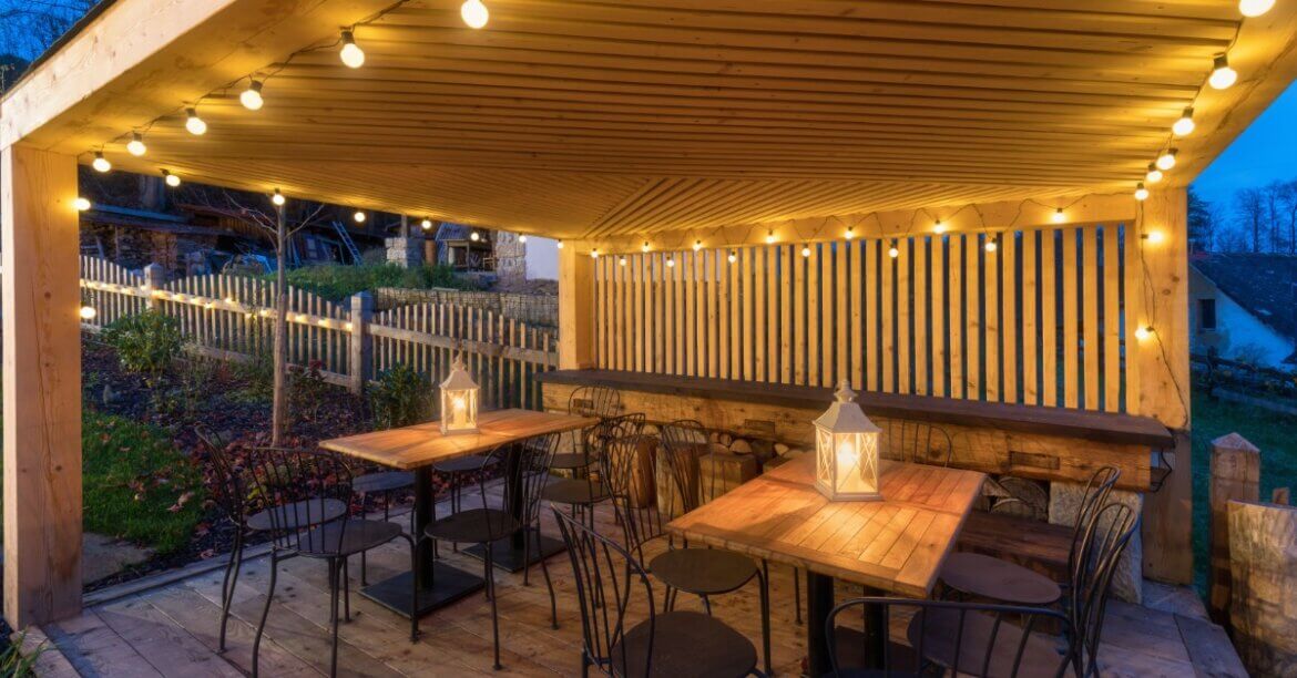 A wooden pavilion with string lights covers several dining tables and chairs in an outdoor gathering space.