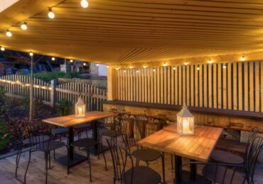 A wooden pavilion with string lights covers several dining tables and chairs in an outdoor gathering space.